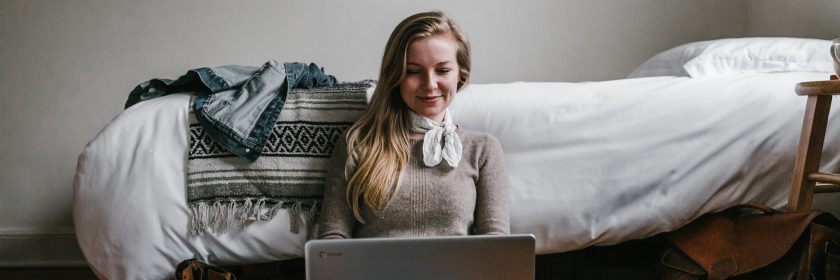 A smiling young woman sits on the floor with her back to a bed, laptop open on her knees.